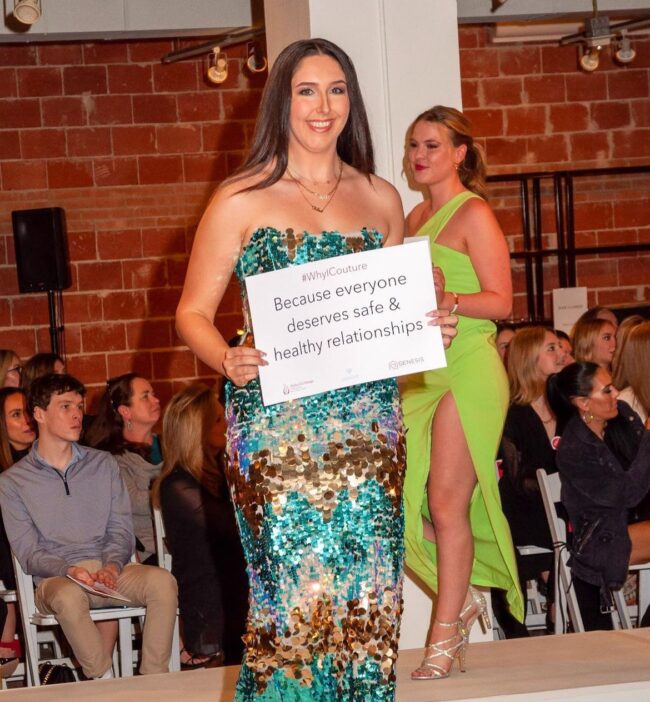 A model sporting fashions from Tootsies walks the runway at Alpha Chi Couture holding a sign representing why she chose to walk in the show to support domestic violence survivors. Image courtesy of SMU Alpha Chi Omega's Instagram.